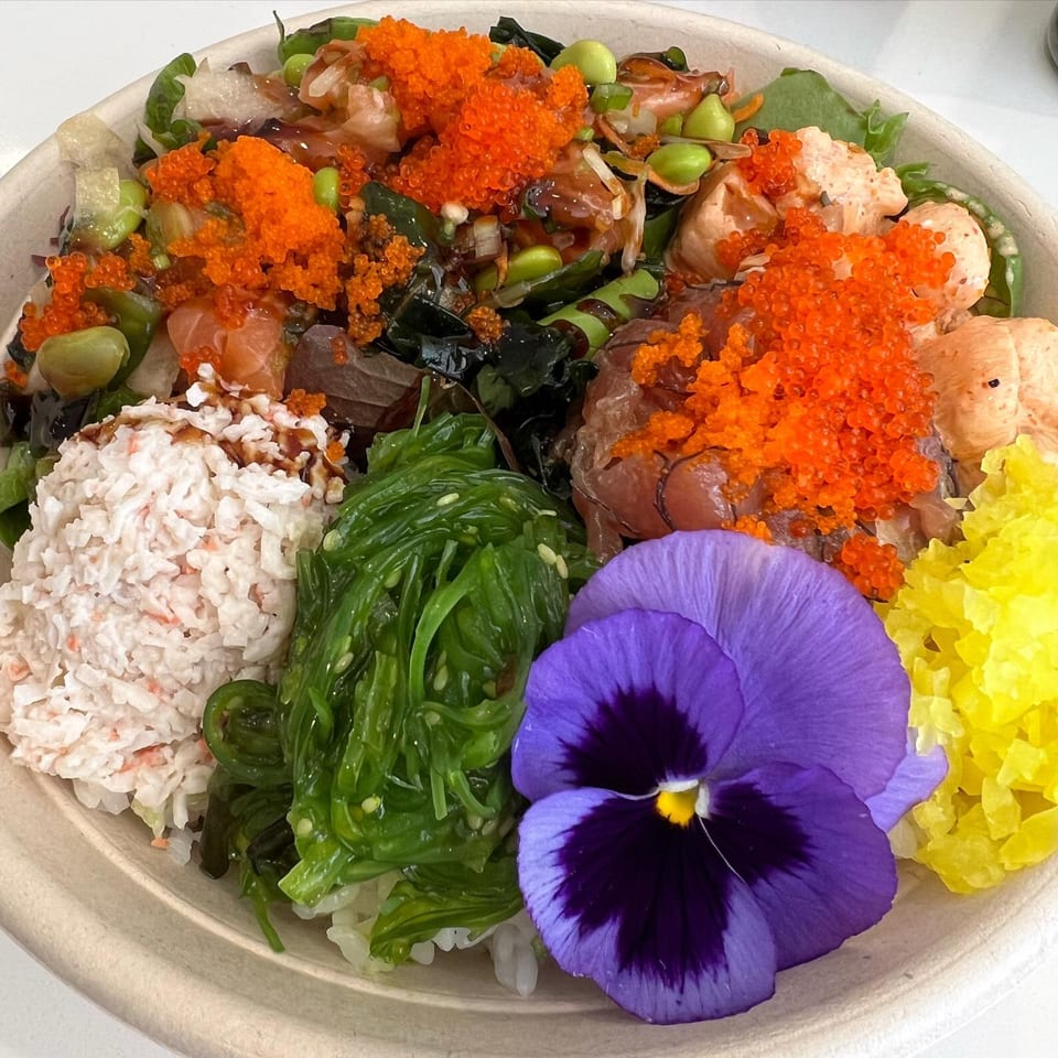 A vibrant Benicia Poke Co. bowl with various toppings, including crab, seaweed, roe, and a purple flower.